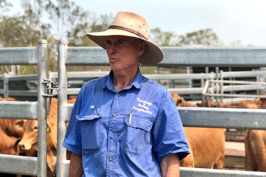 A man in a blue shirt and wide-brimmed hat looks away with cattle yards behind him