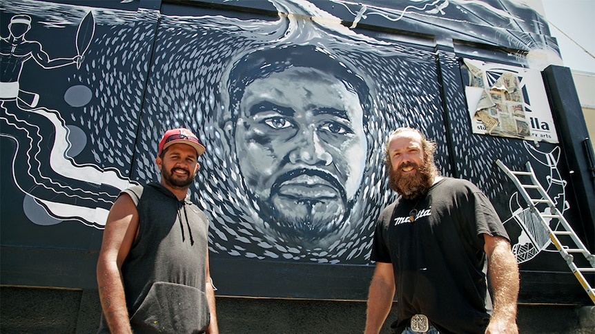 Two men smiling in front of a mural of with Indigenous artwork