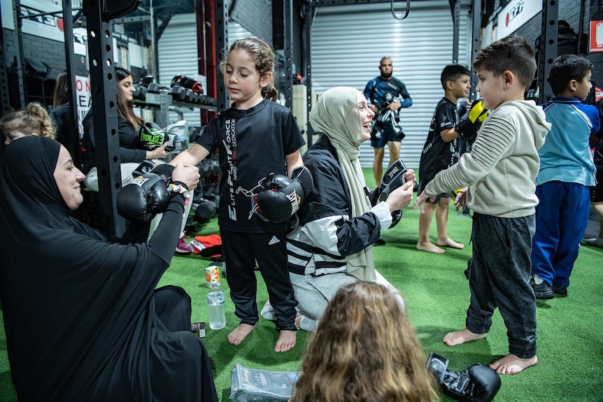 Two mothers smile while helping their children put on boxing gloves.