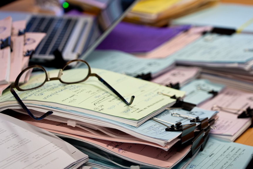 A stack of lawyer's files strewn across a table, with a pair of glasses on top of one pile.
