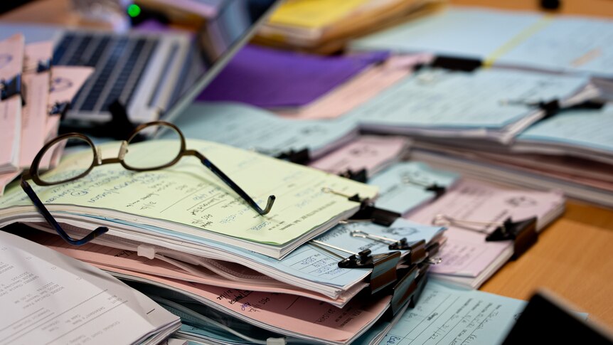 A stack of lawyer's files strewn across a table, with a pair of glasses on top of one pile.