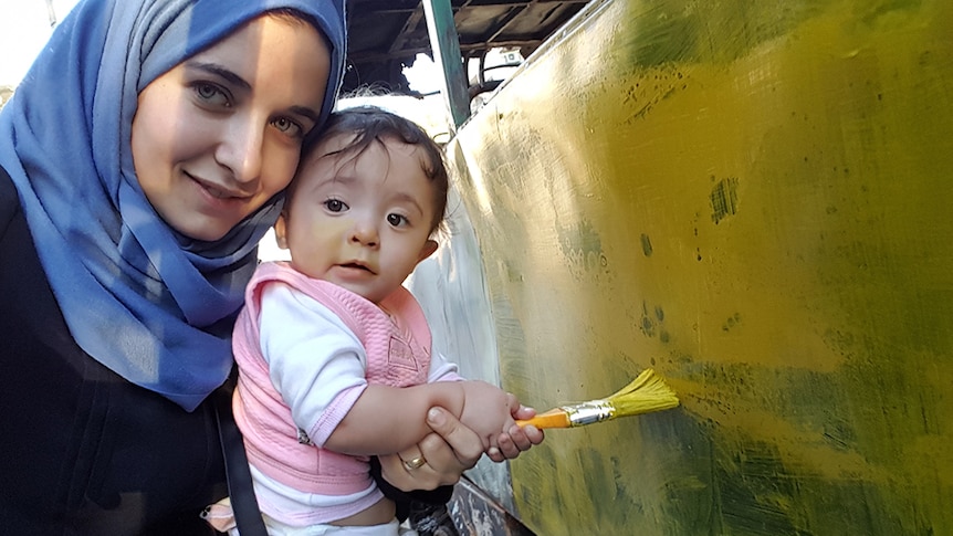 A woman with blue headscarf  holds her young baby daughter, both hold brush and paint an outdoor structure with yellow paint.