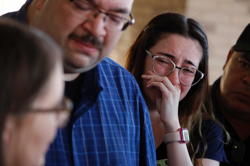 A woman wearing glasses wipes tears away as her parents speak with the media.
