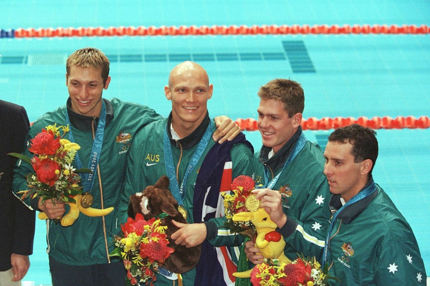 Four Olympic swimmers in team Australia tracksuits hold flowers and wear medals poolside