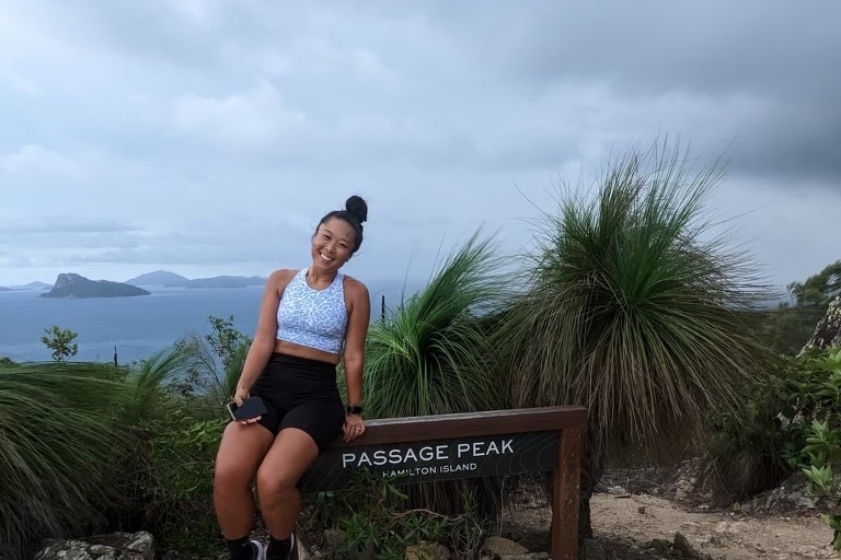 A young woman in activewear sitting on a 'passage peak, Hamilton Island' sign with the blue ocean visible in the background. 