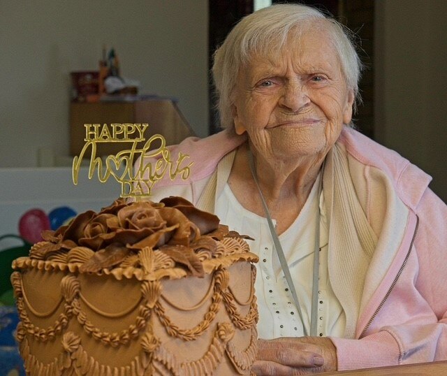 Woman sits in front of cake that reads "Happy Mothers Day" She has white hair, wears pink jumper.