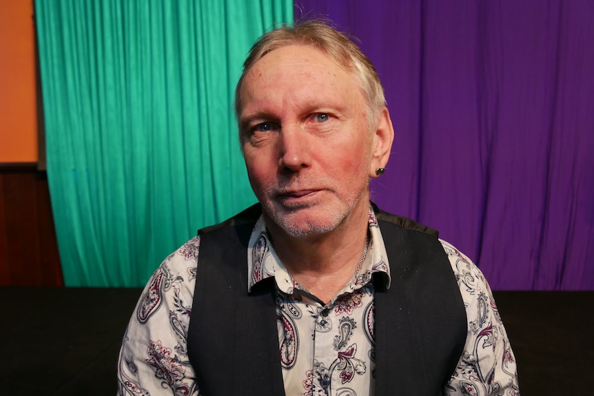 A man in a floral shirt and vest smiles at the camera with green and purple stage curtains in the background 