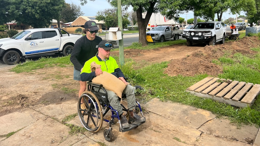Two men, one who is using a wheelchair, carry a sandbag up a driveway