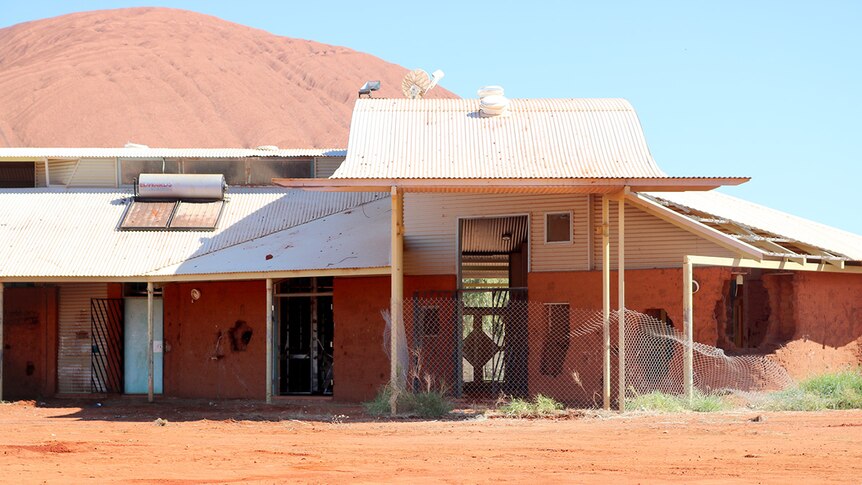 The old adult education centre in Mutitjulu, surrounded by dirt with holes in the walls, sits in front of Uluru.