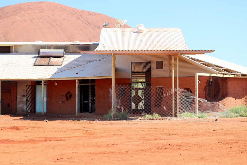 The old adult education centre in Mutitjulu, surrounded by dirt with holes in the walls, sits in front of Uluru.