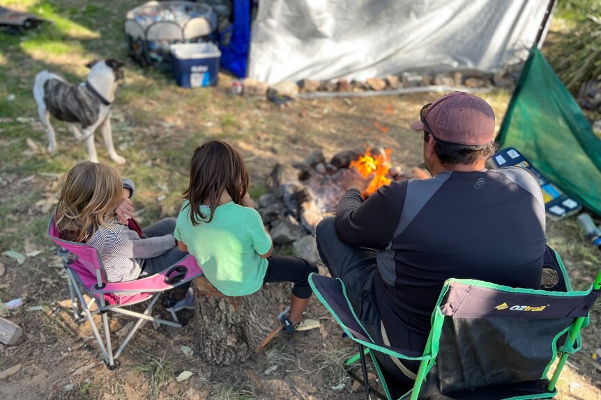 Back of father, two kids and dog sitting by a campfire with their tent in the foreground.