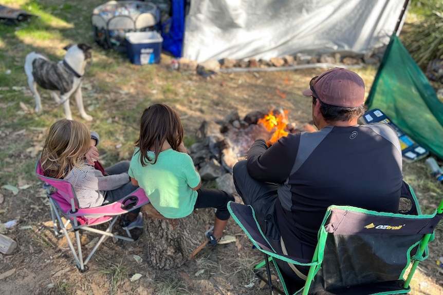 Back of father, two kids and dog sitting by a campfire with their tent in the foreground.