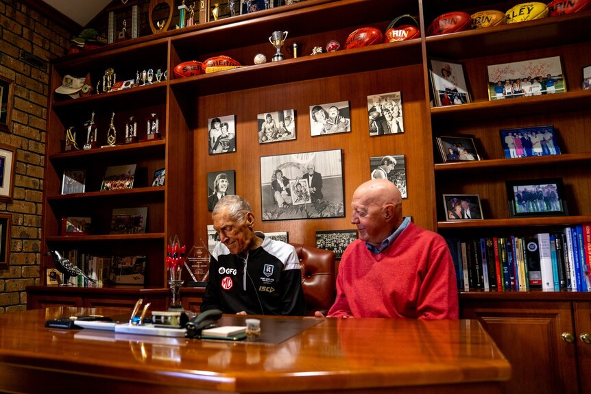 Two old men sit together at a desk surrounded by AFL medals and paraphernalia.
