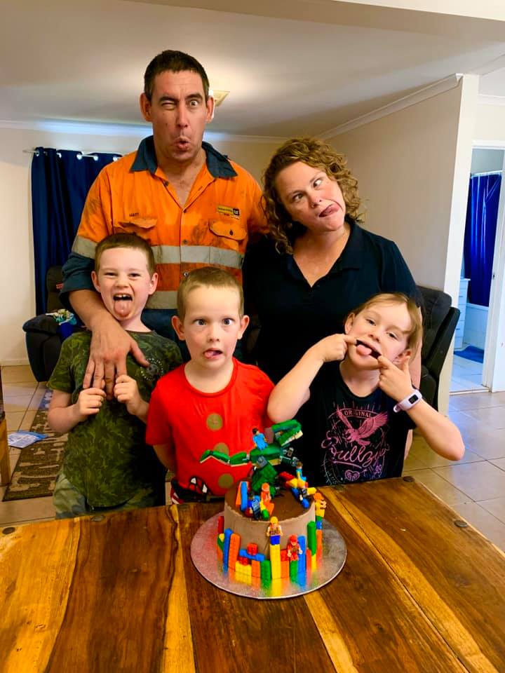 A woman and her husband pose with their three kids behind a kitchen table with a birthday cake