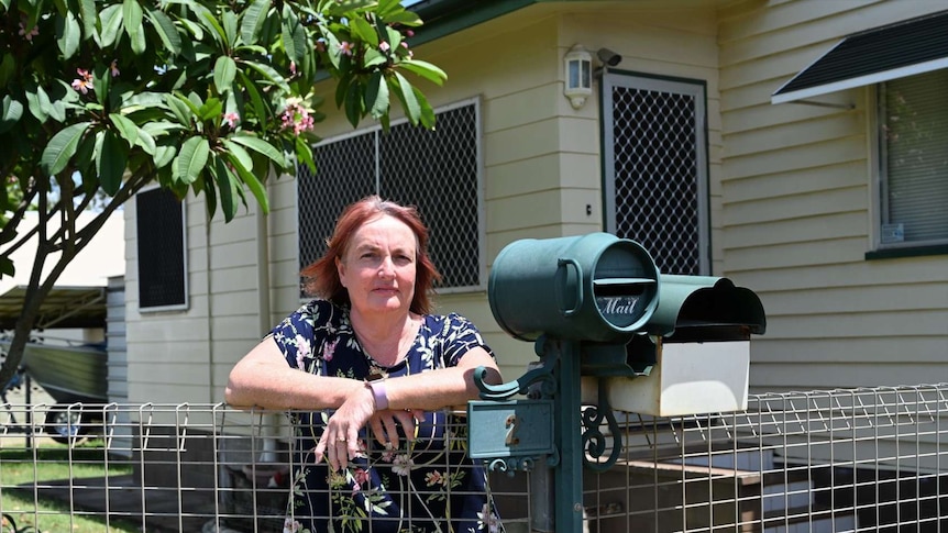 A woman with red hair stands with folded arms leaning on a fence near her letterbox with a frangipani tree in the background.