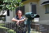 A woman with red hair stands with folded arms leaning on a fence near her letterbox with a frangipani tree in the background.