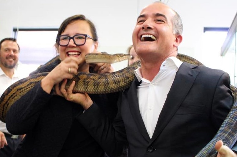A man with a large snake around his neck laughs as a woman next to him smiles.