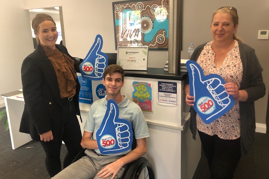 Boy sitting in wheelchair, next to two ladies all holding big blue thumps up signs