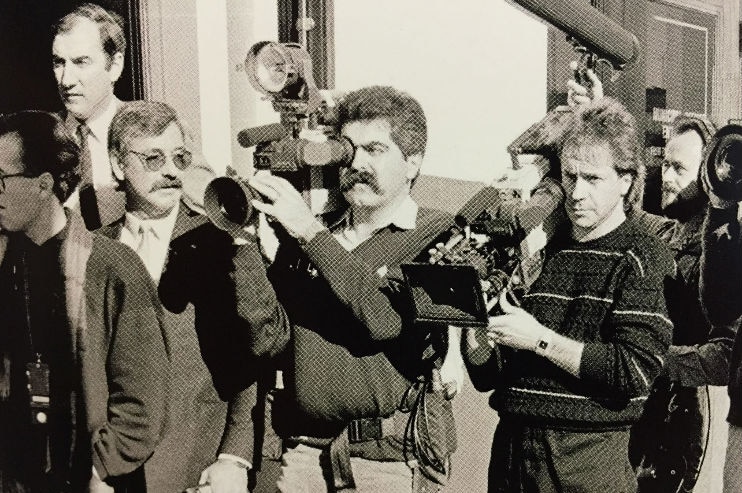 Black and white photo of Vince holding camera amongst media pack.