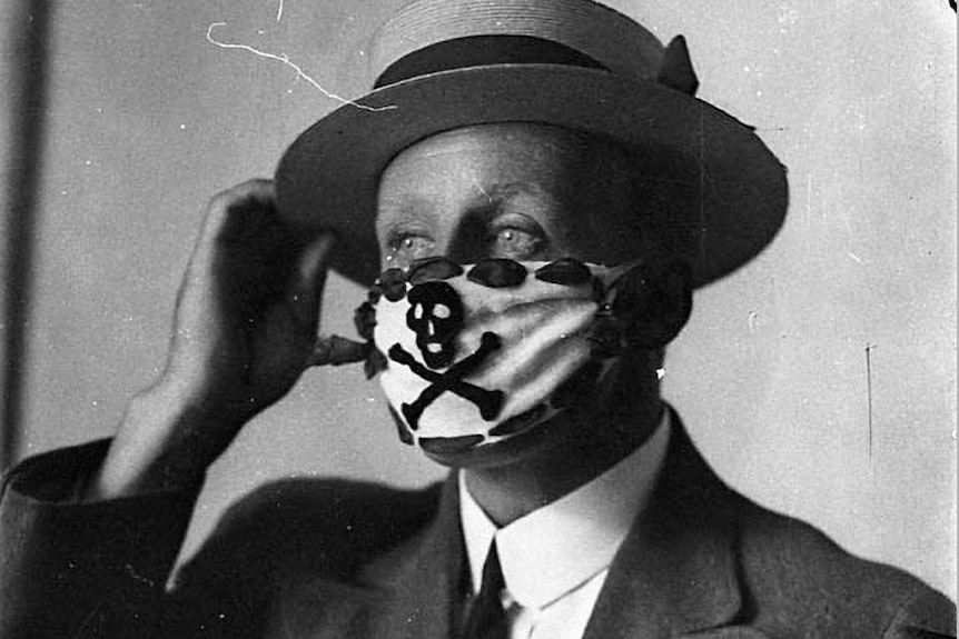 Black and white photo of a man wearing a suit, tie, boater hat and a protective facemask bearing skull and crossbones.