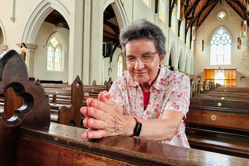 A woman sits in a pew in a cathedral hands folded with her eyes shut