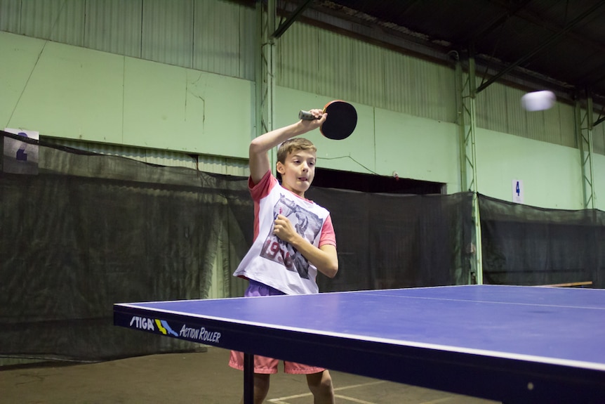 Evan McAllister plays a forehand table tennis shot with a look of concentration on his face in a shed.