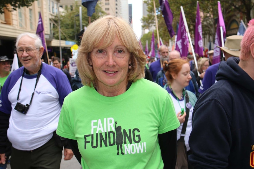 A woman wearing a green t-shirt reading 'Fair Funding Now!' stands amongst a sea of protesters.