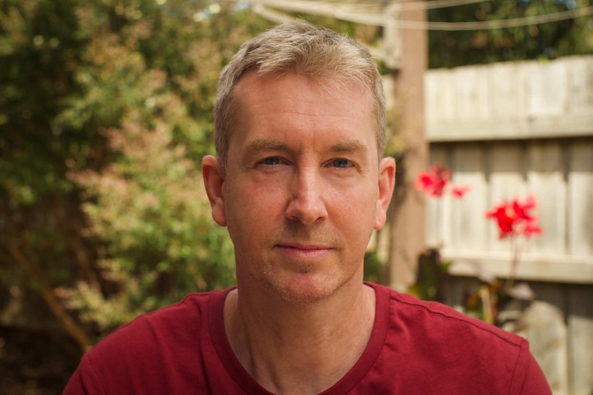 A man with short blond hair stares into the camera as he sits in his garden.