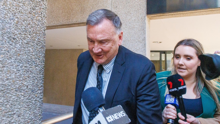 Middle aged man in suit, flanked by reporter, ABC microphone in foreground
