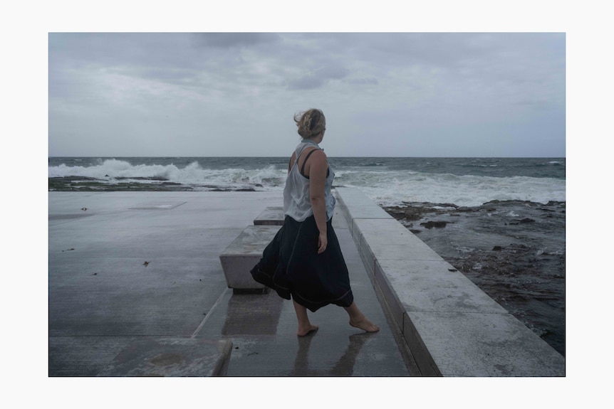 Clementine stands before a grey scene of crashing waves as she walks alongside an ocean pool