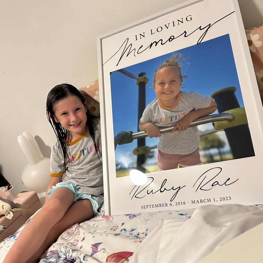a little girl sits on her bed with a large framed photo of her best friend with the words 'in loving memory' on it