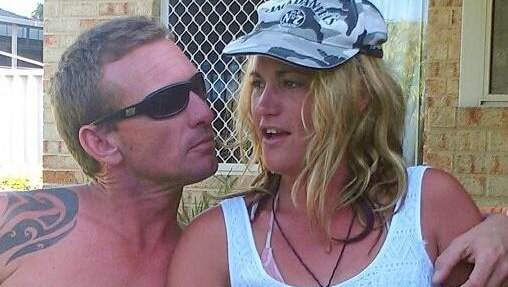 A man with a bare chest and sunglasses sits with his arm around a woman in a white singlet and Jack Daniels cap in a garden.