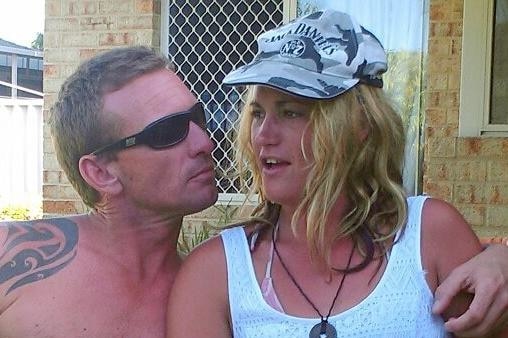 A man with a bare chest and sunglasses sits with his arm around a woman in a white singlet and Jack Daniels cap in a garden.