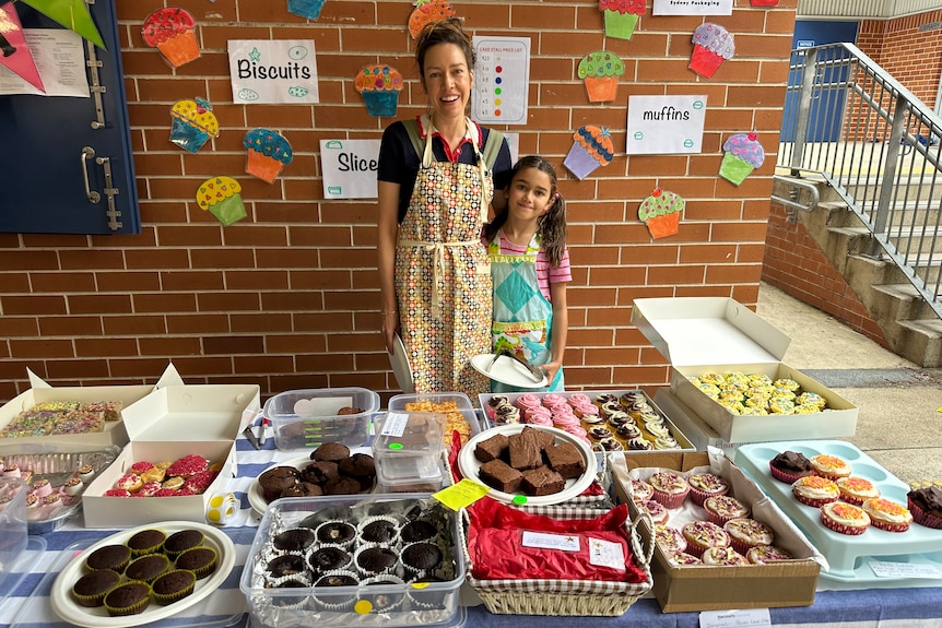 A woman and girl stand at a cake stall in a school