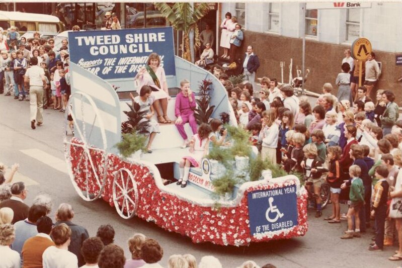 A woman wearing a sash sits on a float which is passing though a crowd watching on a street.