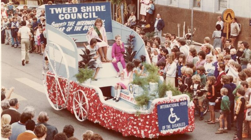 A woman wearing a sash sits on a float which is passing though a crowd watching on a street.