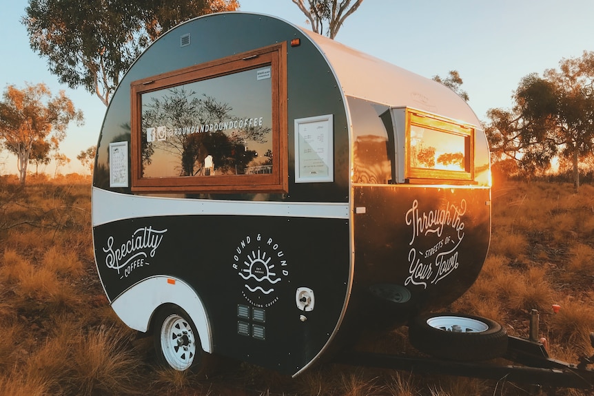 A vintage-looking caravan sits on an outback field, with gum trees around in the late afternoon sunshine.