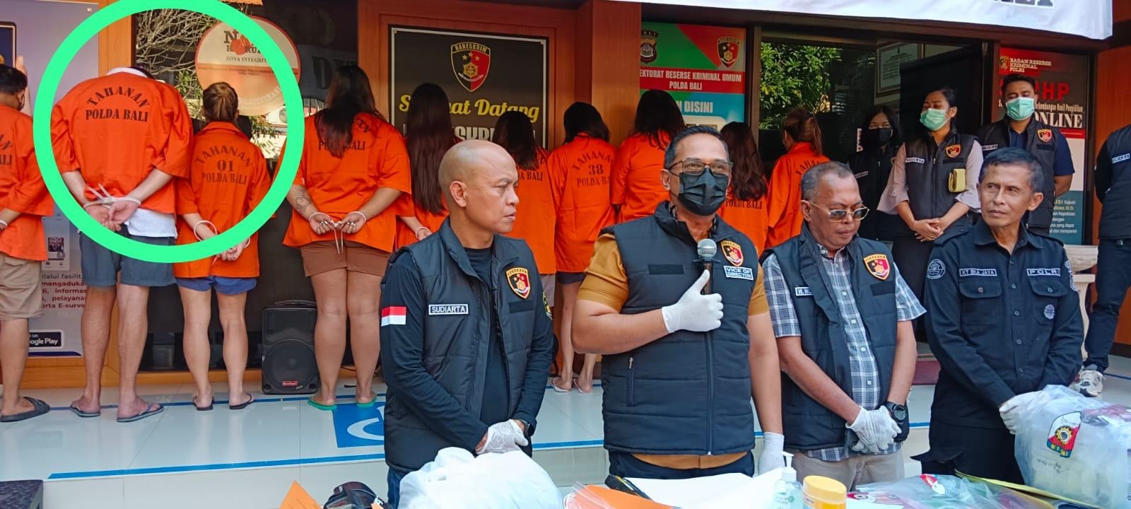 Empat polisi Indonesia berdiri di belakang meja berisi barang-barang dalam kantong plastik. Di belakang mereka, orang-orang berkemeja oranye berdiri berjajar.