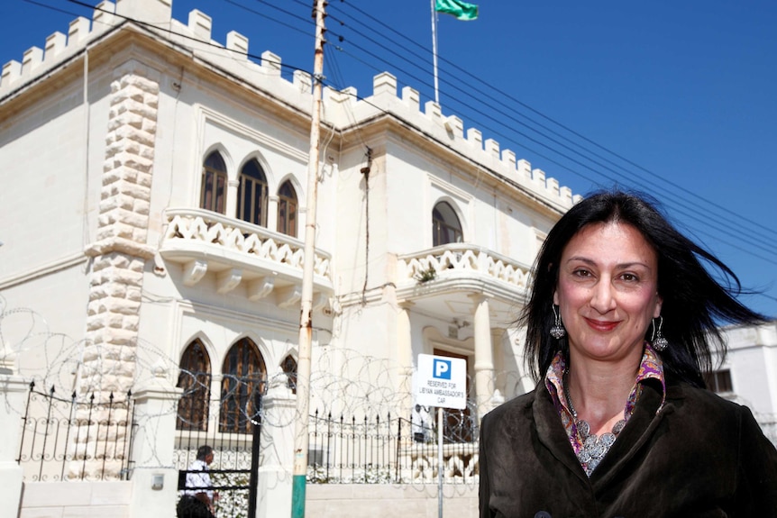 Maltese investigative journalist Daphne Caruana Galizia poses outside a white building with a blue sky background.