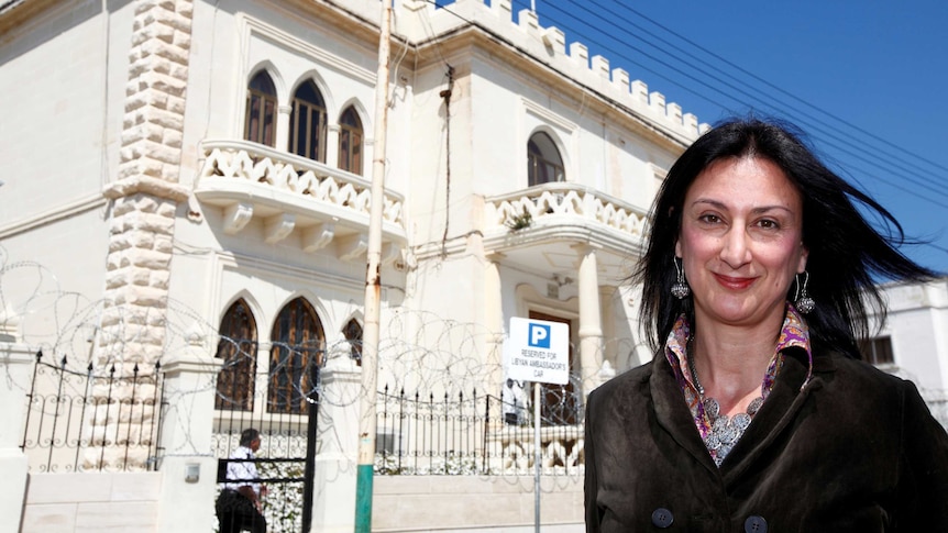 Maltese investigative journalist Daphne Caruana Galizia poses outside a white building with a blue sky background.