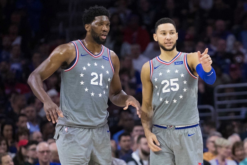 Ben Simmons and Joel Embiid converse on the court. Simmons points to something.