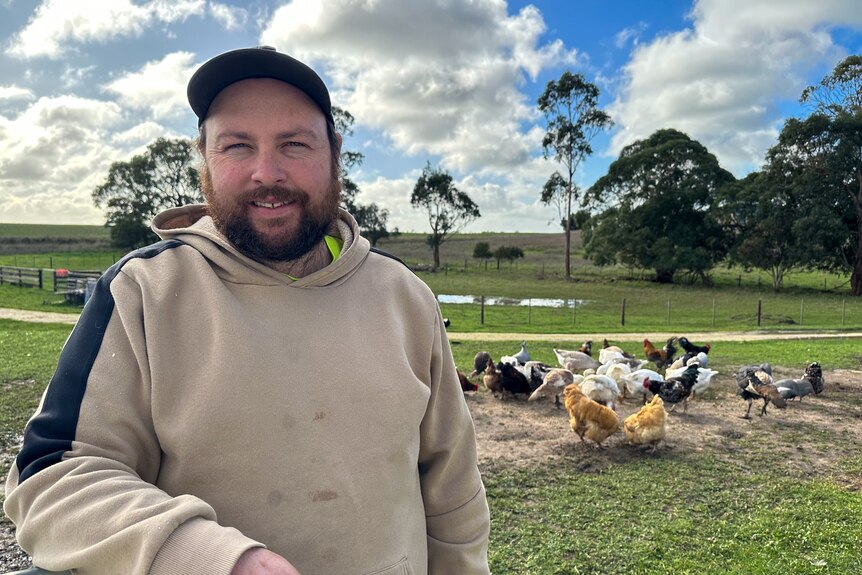 A man wears a cap and smiles at the camera, chickens and ducks can be seen in the background.