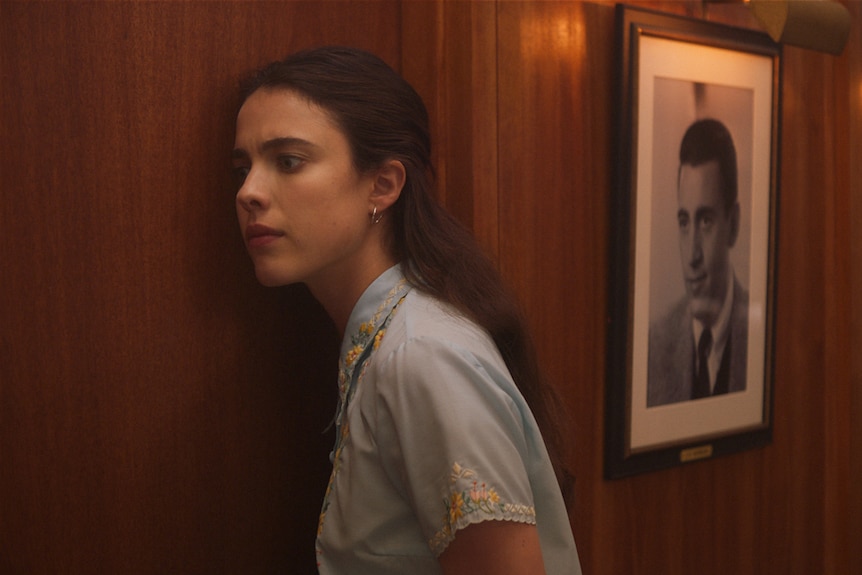 Young woman with long dark brown hair leaning up against a wood-panelled wall with photographic portraits, as if listening.