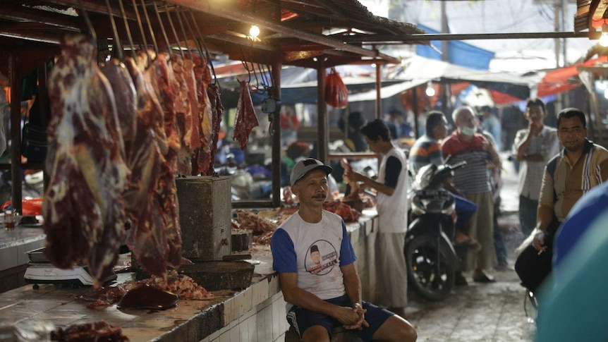 A man in a baseball cap sits at an open butchers stall at a wet market.