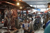 A man in a baseball cap sits at an open butcher's stall at a wet market.