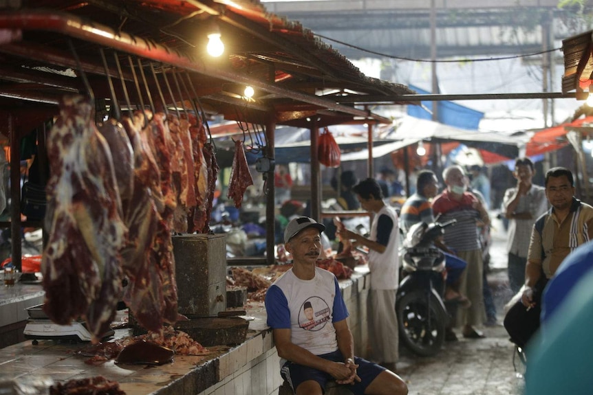 A man in a baseball cap sits at an open butchers stall at a wet market.