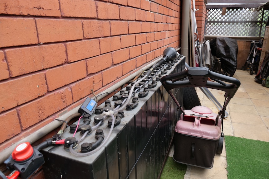 A row of large batteries connected by cables down by the side of a red-bricked wall