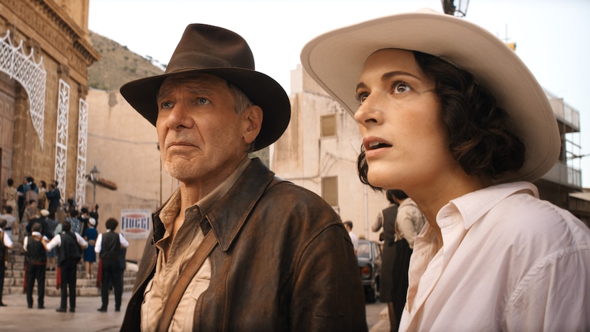 An octogenarian white man in a brown hat and jacket stands in a town square with a white brunette woman in a white hat and shirt