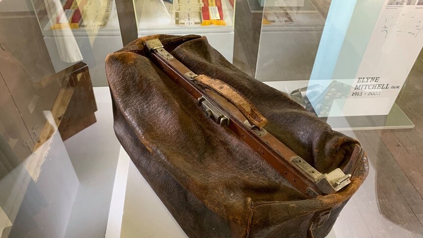 An old leather bag sits in a display case. It is brown and very worn.