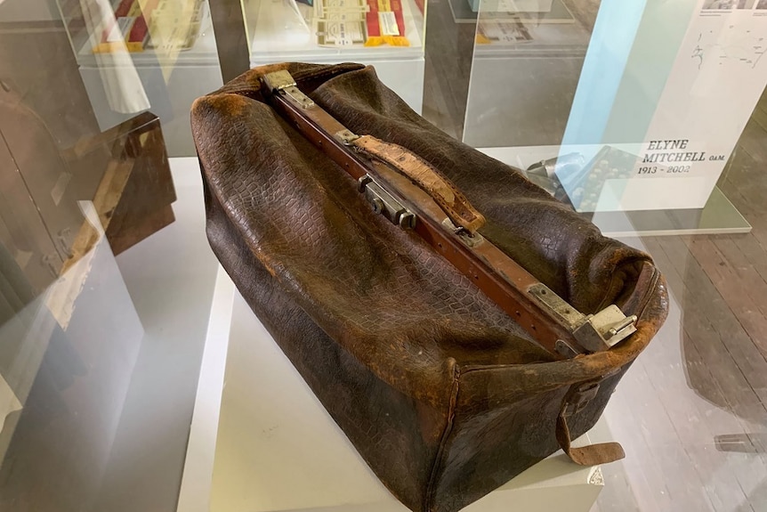 An old leather bag sits in a display case. It is brown and very worn.